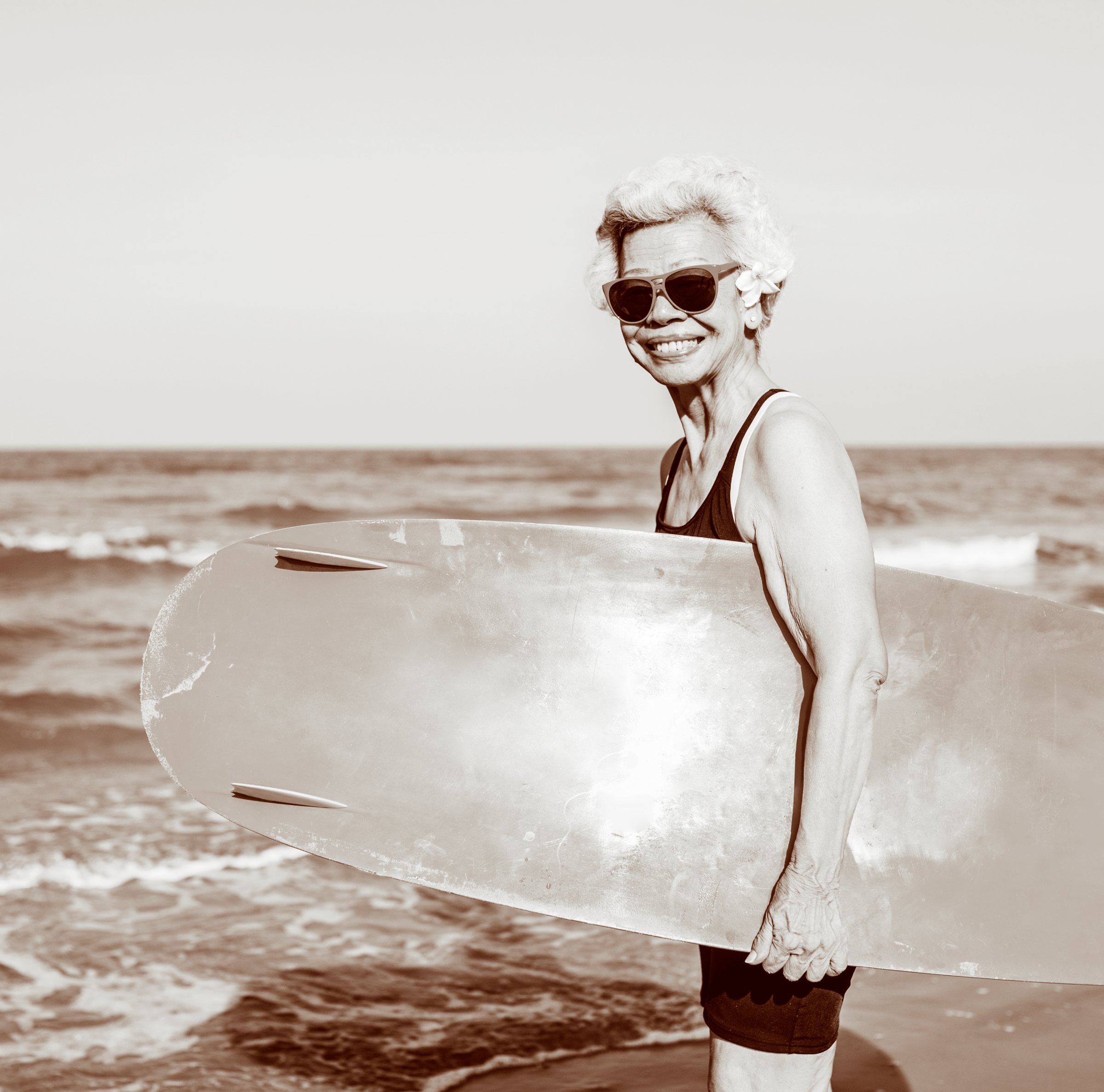 Ältere Frau mit Sonnenbrille steht mit Surfbrett am Strand